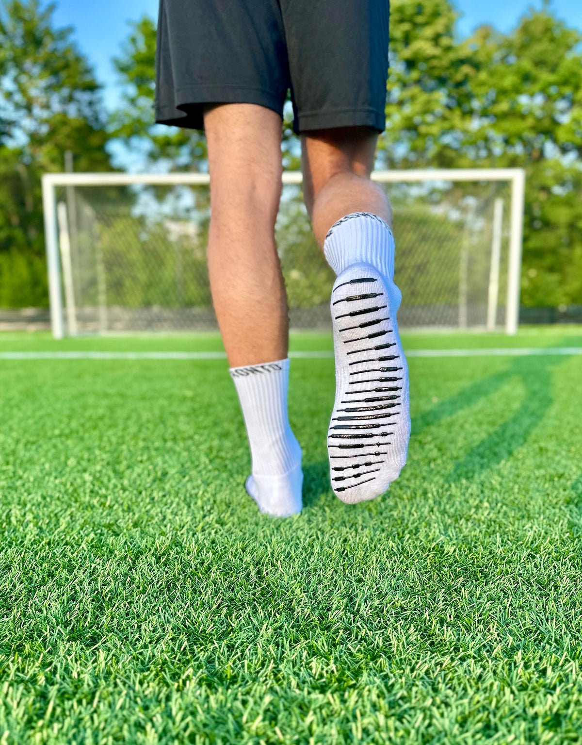 Fodboldspiller iført hvide Pronto-gripsocks går på en grøn fodboldbane med et mål i baggrunden.