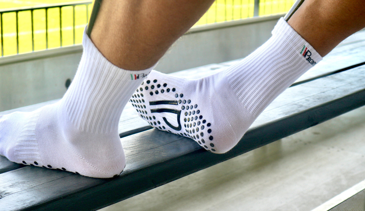 Fødder iført hvide Pronto gripsocks med sorte grebsmønstre, placeret på en bænk med udsigt til en fodboldbane.