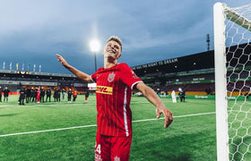 Lucas Høgsberg fra FC Nordsjælland i rød spillerdragt fejrer på banen med armene spredt. Baggrunden viser stadionet med lysmaster og banneret "The Right to Dream".