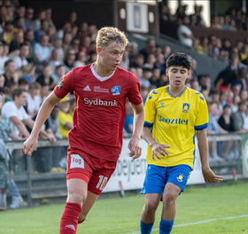 Mathias Kaarsbo i rød spillertrøje under en fodboldkamp mod Brøndby IF, iført gule og blå trøjer, foran publikum
